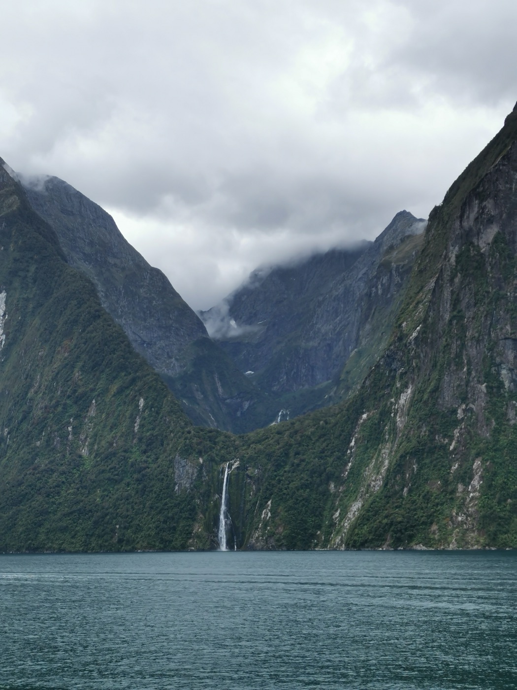 A tranquil fiord with a waterfall between steep forested cliffs.