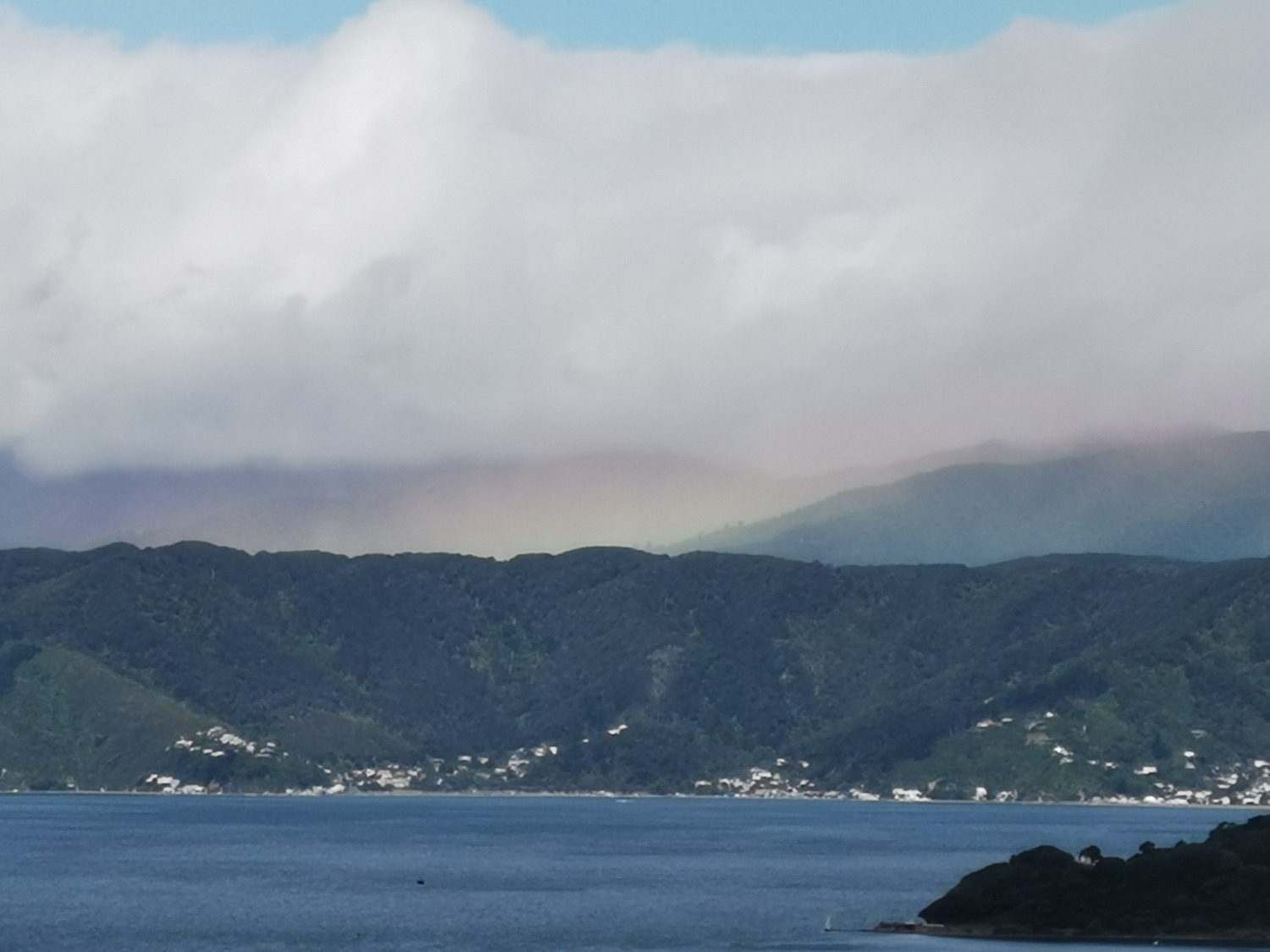 Soft light and a faint rainbow over a calm harbour and green hills.